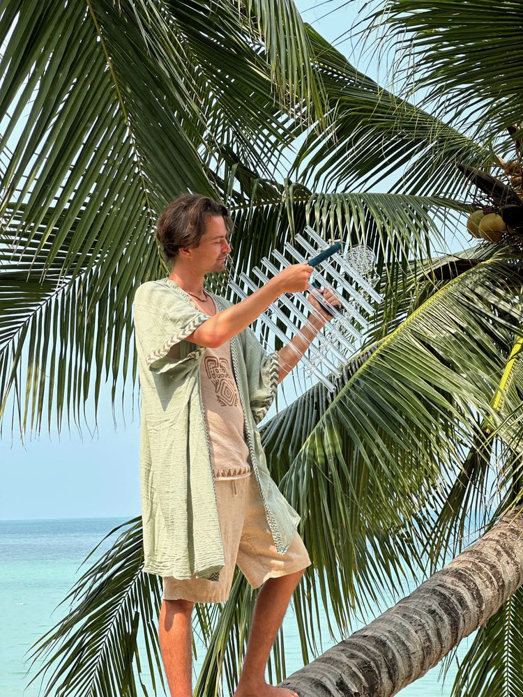 Young man playing a crystal harp while standing on a palm tree trunk, tropical blue sea and coconut palms in the background