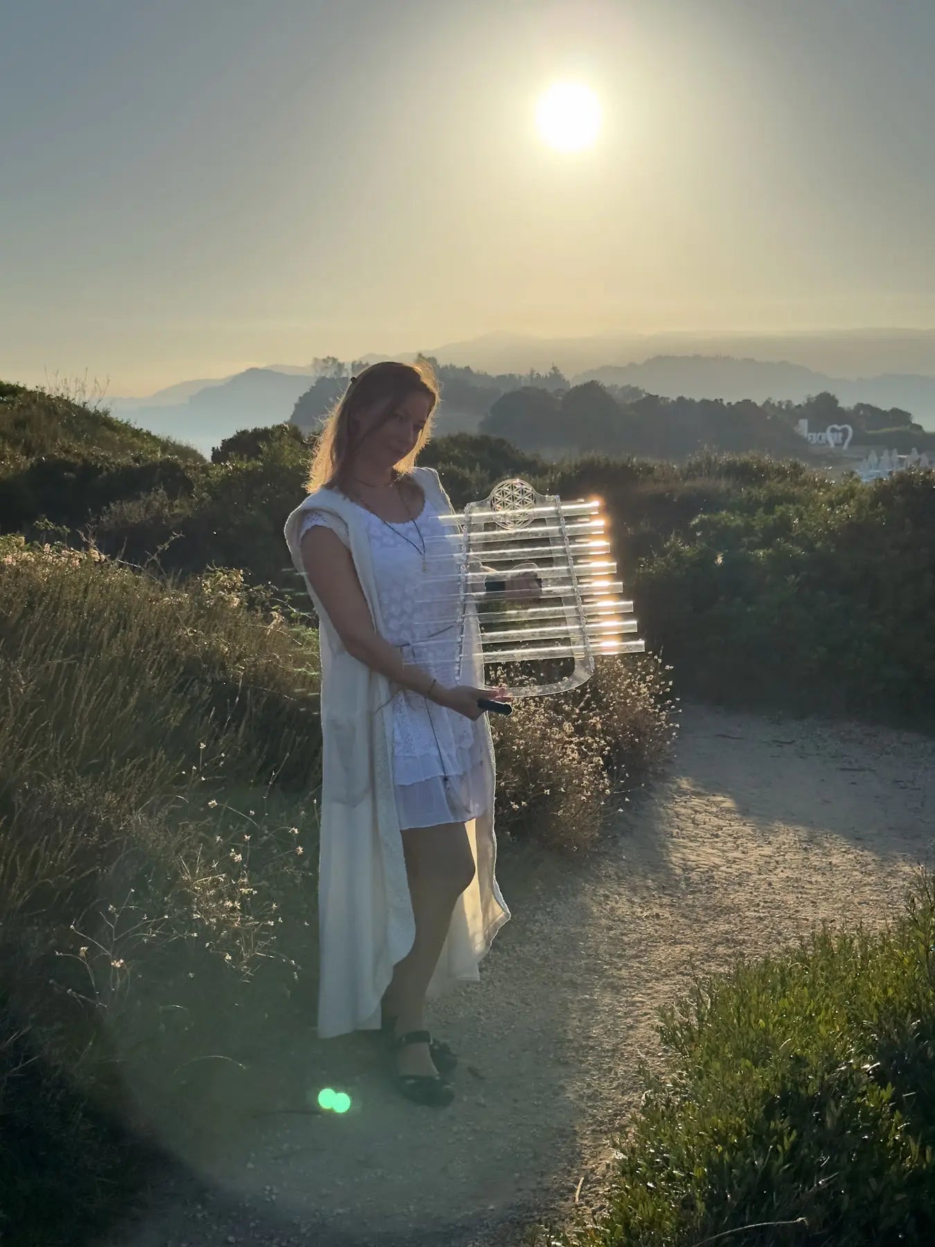 Woman in white dress holding a crystal harp on a sandy hilltop path at sunset, sun glowing behind her with misty mountains in the distance