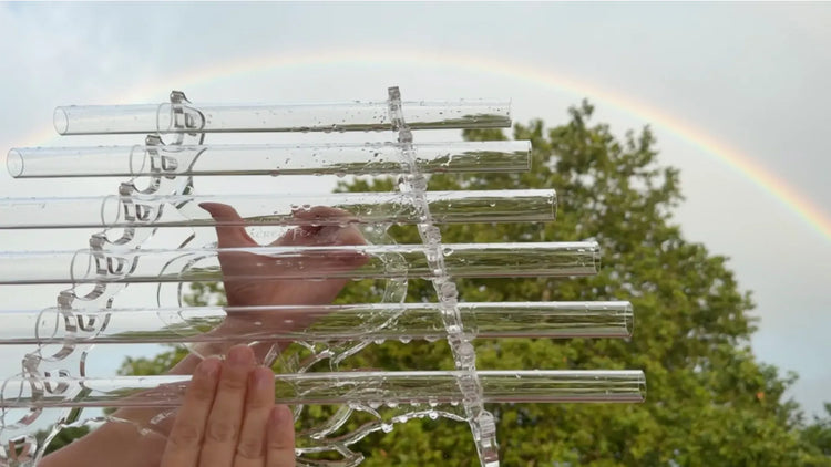 Wet hands playing a crystal harp with water droplets on quartz tubes, rainbow arching across sky behind green trees